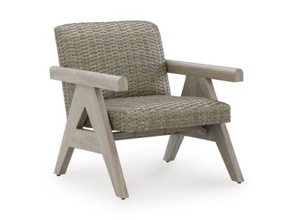 A single, stylish outdoor armchair photographed against a plain, white background. The chair has a modern, slightly rustic design that combines a light wood frame with a woven seat and backrest. The wooden frame appears to be made from a pale, natural-toned wood with a smooth, matte finish. Its structure is sturdy and geometric, featuring angled legs that splay outward slightly for stability. The armrests are thick and straight, extending horizontally from the back toward the front. Each armrest is supported by a distinctive triangular side structure formed by the legs and cross supports, giving the chair a sculptural, architectural look. The front legs angle forward while the rear legs angle slightly backward, creating a balanced stance. The seat and backrest are made of tightly woven material in a neutral beige or taupe color, resembling wicker or rattan. The weaving pattern is consistent and textured, adding visual interest and a tactile quality. The backrest is slightly reclined, suggesting comfort, while the seat is firm and gently contoured. Small, dark protective caps are visible at the bottom of each leg, likely to prevent floor damage. Overall, the chair has a clean, contemporary aesthetic with natural materials and a neutral color palette, making it suitable for both indoor and outdoor settings.